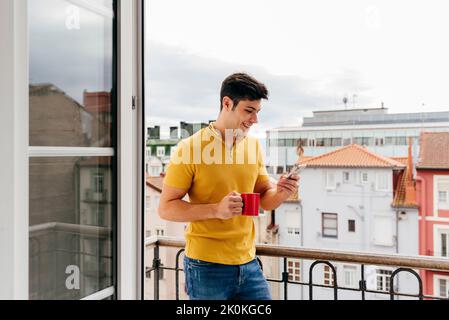 Attractive caucasian man with a coffee cup on the balcony at home using mobile phone Stock Photo