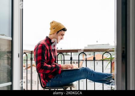 Side view of trendy focused caucasian man with beanie hat and shirt sitting on chair at balcony while working remotely on laptop at home Stock Photo