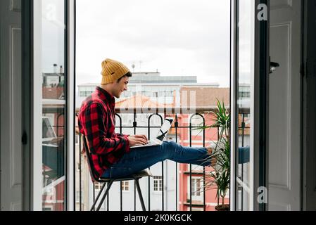 Side view of trendy focused caucasian man with beanie hat and shirt sitting on chair at balcony while working remotely on laptop at home Stock Photo