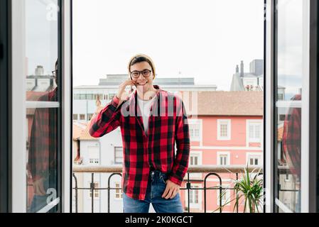 Attractive cheerful hipster caucasian man with beanie hat an glasses standing on balcony at home speaking on mobile phone Stock Photo