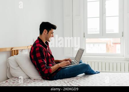 Side view of handsome caucasian man working on the laptop in bedroom at home Stock Photo