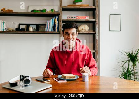 Handsome happy caucasian man eating at home while having lunch break from working with laptop on the table looking at camera Stock Photo