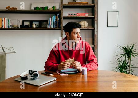 Handsome thoughtful caucasian man eating at home while having lunch break from working with laptop on the table Stock Photo