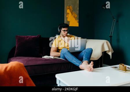 Attractive caucasian man sitting on the couch listening music on headphones and working with the laptop at home Stock Photo