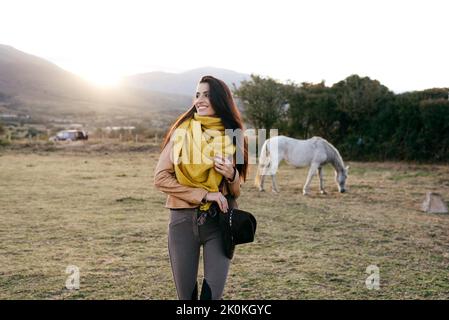 Cheerful woman in scarf walking on ranch field with pasturing horses in sunlight on background Stock Photo