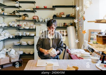 Focused mature female master polishing and smoothing clay cup with professional tool while creating pottery in craft workshop Stock Photo