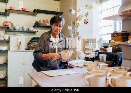 Focused mature female master polishing and smoothing clay cup with professional tool while creating pottery in craft workshop Stock Photo