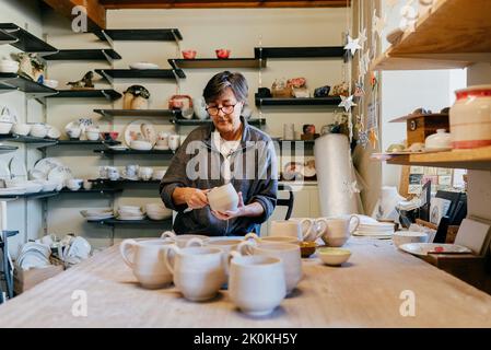 Focused mature female master polishing and smoothing clay cup with professional tool while creating pottery in craft workshop Stock Photo
