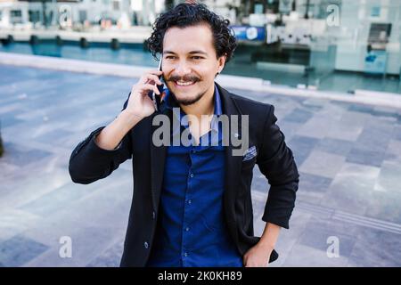 Content bearded male entrepreneur in elegant outfit having phone conversation and looking away while standing on pavement near building on street Stock Photo