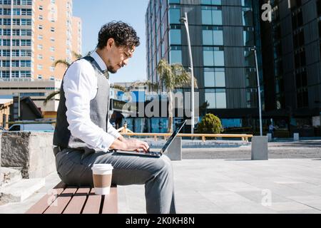 Side view of focused bearded male entrepreneur in elegant suit browsing netbook while sitting on bench with takeaway coffee on sunny street Stock Photo