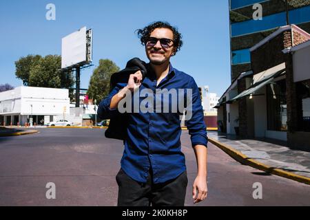 Positive bearded male in sunglasses wearing trendy shirt smiling and strolling with jacket in hand on sunny street with buildings Stock Photo