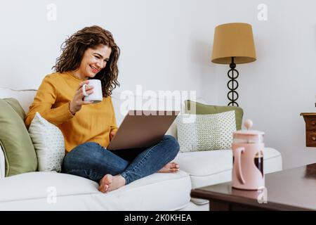 Positive woman freelancer in casual clothes sitting with crossed legs on comfortable sofa and drinking cup of tea while using netbook in living room a Stock Photo