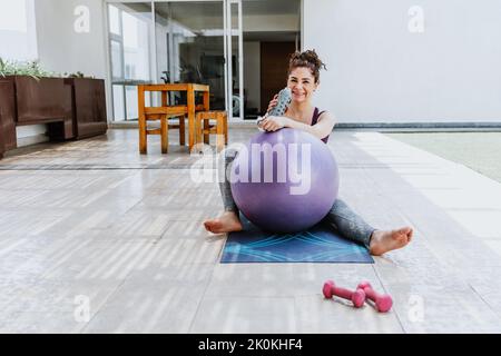 Full body of cheerful young barefooted female in sportswear sitting on mat with fit ball and smiling while drinking bottle of water after workout Stock Photo
