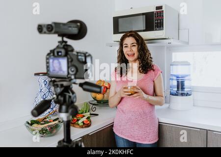 Positive female on casual clothes standing in kitchen and looking at camera on tripod while recording video for vlog about healthy food Stock Photo