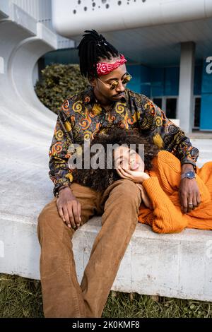 Happy black man sitting and woman lying down in stylish casual outfits cuddling on concrete bench Stock Photo