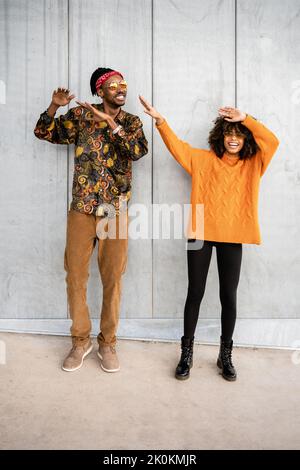Cheerful stylish African American woman and man in bright clothes dancing and laughing against gray street wall Stock Photo