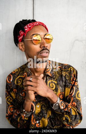 Thoughtful African American guy with dreadlocks wearing stylish round sunglasses with red headband and colorful shirt looking away Stock Photo