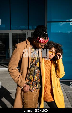 African American man and woman in stylish beige and yellow coats embracing each other while walking on the on street looking away Stock Photo