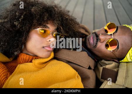 Top view of trendy African American man and woman in bright coat lying down on wooden floor looking at each other Stock Photo