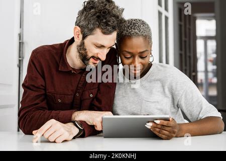 Satisfied African American woman and bearded man smiling while sitting at table and watching digital tablet together at home Stock Photo