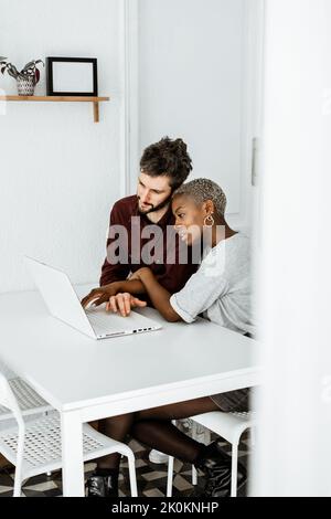 Side view from above of contemporary African American woman sitting at table and working with laptop with young man surfing digital table at home Stock Photo