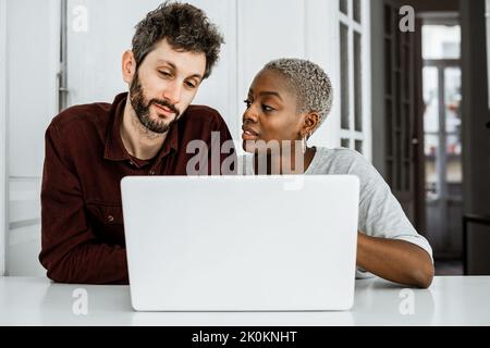 Contemporary African American woman sitting at table and working with laptop with young man surfing digital table at home Stock Photo
