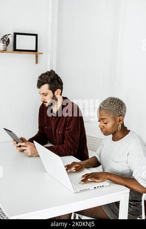 Side view from above of contemporary African American woman sitting at table and working with laptop with young man surfing digital table at home Stock Photo