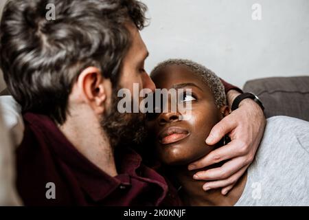 Young bearded man in casual shirt lying on comfortable couch in embrace with African American woman in cozy living room looking at each other Stock Photo