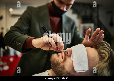 Crop male barber in mask using razor and shaving beard of client in barbershop Stock Photo