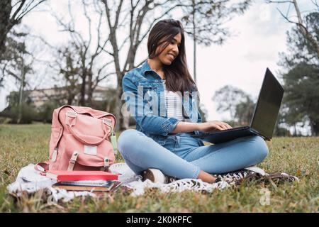 Smiling ethnic female university student with backpack and books sitting on grass and doing homework on laptop while spending time in park Stock Photo