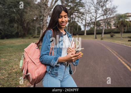 Happy young ethnic female student with backpack and books using mobile phone and looking at screen while strolling on asphalt path in green park Stock Photo