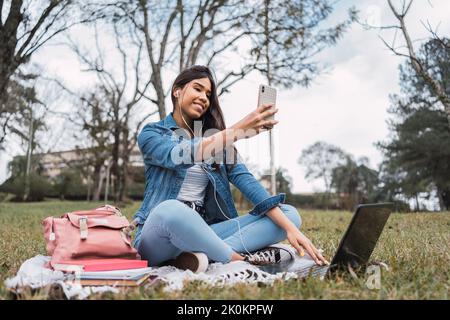 Positive young Hispanic female student in denim outfit taking selfie on smartphone to share with friends in social networks while studying online with Stock Photo