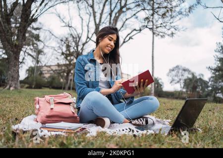 Positive young Hispanic female student in denim clothes with laptop and earphones listening to music and reading book while doing homework assignment Stock Photo