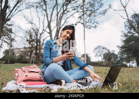 Positive young Hispanic female student in denim outfit browsing on smartphone while studying online with laptop and books in park Stock Photo