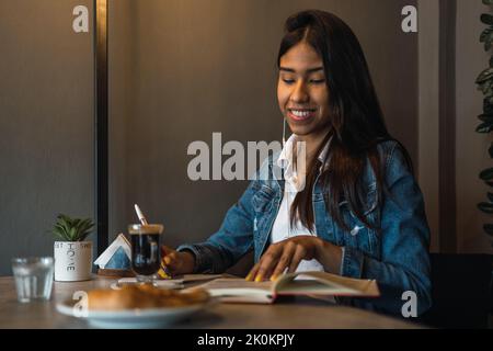 Content smart young Hispanic female student writing in copybook while doing homework assignment at table drinking coffee Stock Photo