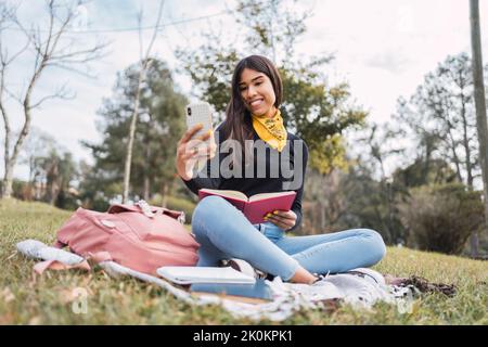 Cheerful young Hispanic female student making video call on mobile phone while studying with open book on green meadow in park Stock Photo