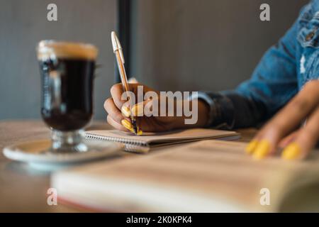 Cropped unrecognizable hispanic female student writing in copybook while doing homework assignment at table drinking coffee Stock Photo