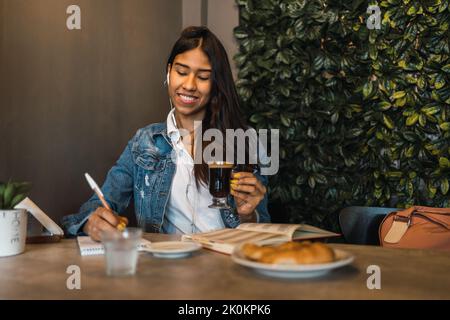 Content smart young Hispanic female student writing in copybook while doing homework assignment at table drinking coffee Stock Photo