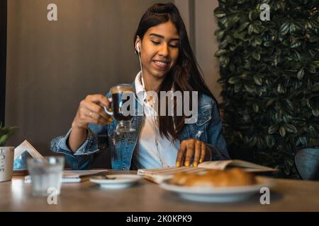 Happy young Hispanic female university student drinking coffee and listening to music with earphones while reading book during exam preparation in caf Stock Photo