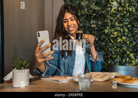Happy young Hispanic female student with coffee in hand taking selfie on smartphone while having coffee break during studies at table with book and no Stock Photo