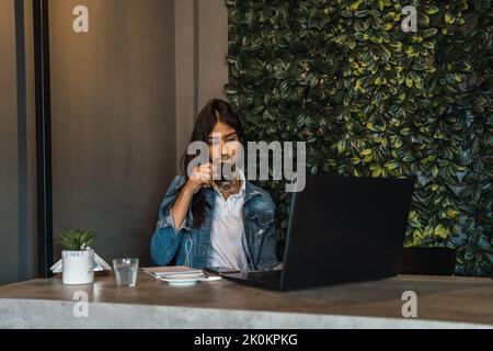 Young Hispanic female freelancer drinking coffee and using laptop while working on remote project at table in cafe Stock Photo