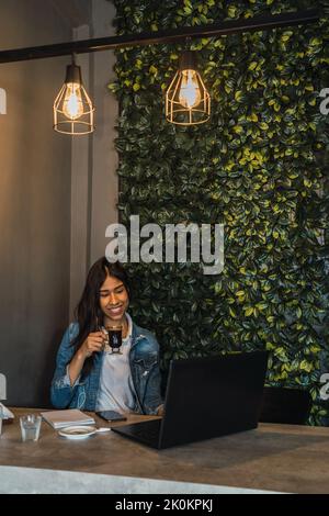 Young Hispanic female freelancer drinking coffee and using laptop while working on remote project at table in cafe Stock Photo