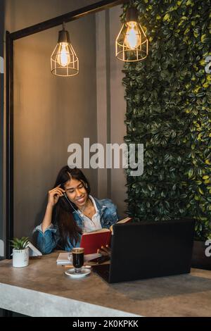 Focused young Hispanic female university student listening to music through earphones and reading book while sitting at table with cup of coffee and l Stock Photo