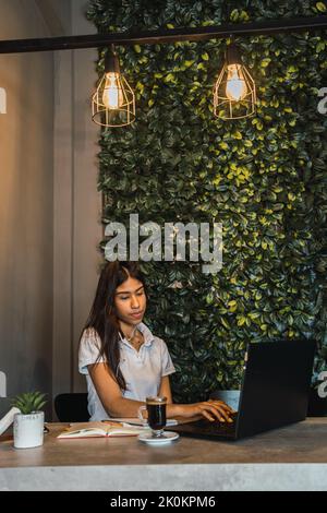 Young Hispanic female freelancer using laptop while working remotely at table with cup of coffee and notebook in cafe Stock Photo