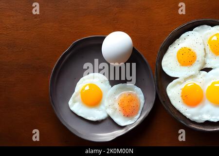 A top view of fried eggs and a raw egg on a black plate against a wooden background. Stock Photo
