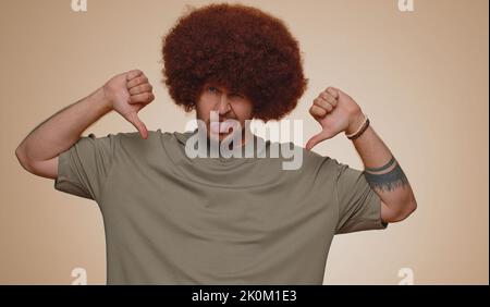 Dislike. Upset unhappy handsome man in t-shirt showing thumbs down sign gesture, expressing discontent, disapproval, dissatisfied, dislike. Young adult guy. Indoor studio shot on beige background Stock Photo