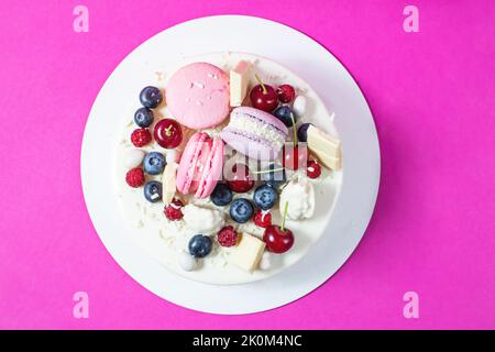 Tender white cake decorated with melted white chocolate, macaroons, meringues, berries and candies on pink background. Top view Stock Photo
