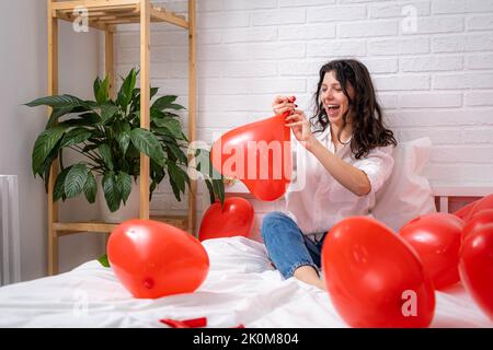 Girl is blowing air balloon at home Handsome brunette girl inflating red heart shaped balloon sitting on bed. Preparation for valentines day party Stock Photo