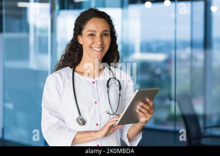 Portrait of young beautiful female doctor with tablet computer, Hispanic female doctor working in modern clinic office smiling and looking at camera wearing white medical coat and stethoscope. Stock Photo