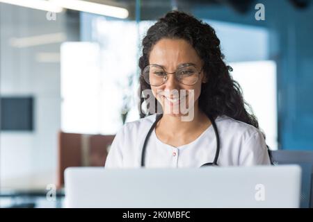Close-up photo of hispanic female doctor working on laptop smiling and happy, latin american woman in medical gown looking at laptop screen working inside modern clinic office Stock Photo